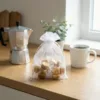 Witte organza zakje 15x20 cm gevuld met fudge snoepjes op een houten blad naast een koffiepot.